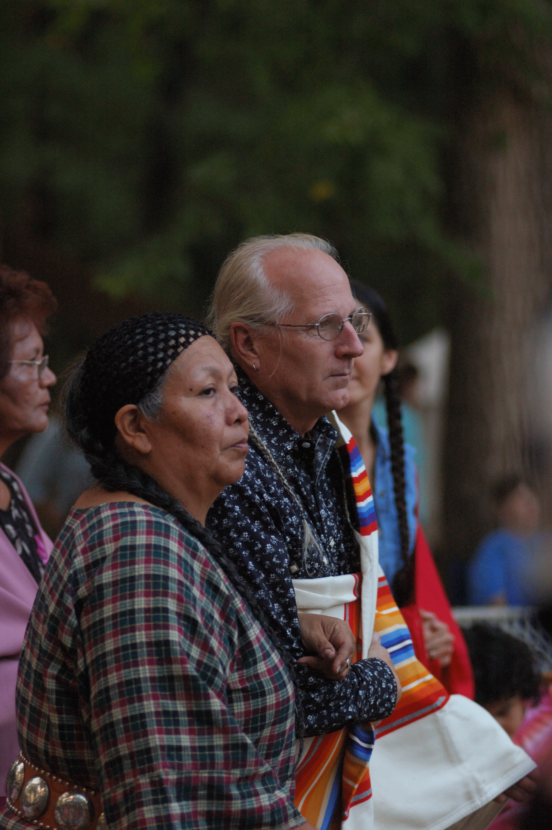 Photo of an older couple in profile. She wears long dark braids, a bandana, and a plaid shirt. He has glasses and white hair and wears a colorful striped textile over his shoulder.