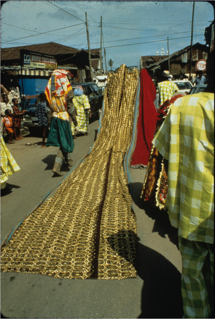 Some Egúngún combine exquisite cloth ensembles with elaborately carved headdresses, as seen in this photo from 1978. This headdress, from a hunter’s family, is distinguished by its dramatic coiffure, a long tuft of hair that descends to the left and terminates with a gourd amulet.